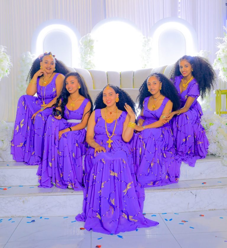 Cinq jeunes femmes en robes violettes posent debout devant un mur de fleurs blanches