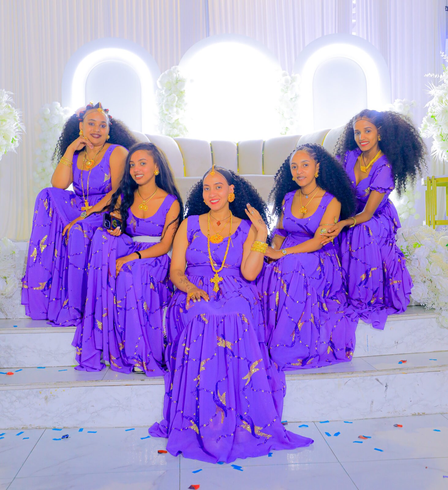 Cinq jeunes femmes en robes violettes posent debout devant un mur de fleurs blanches