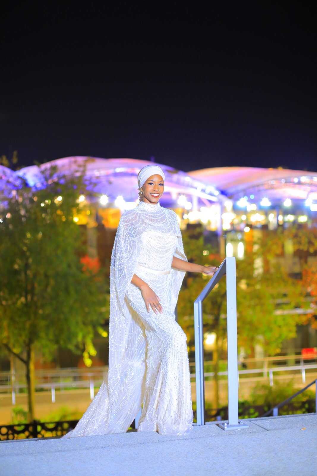 Une femme en robe blanche brodée accoudée à une rambarde, lumières nocturnes