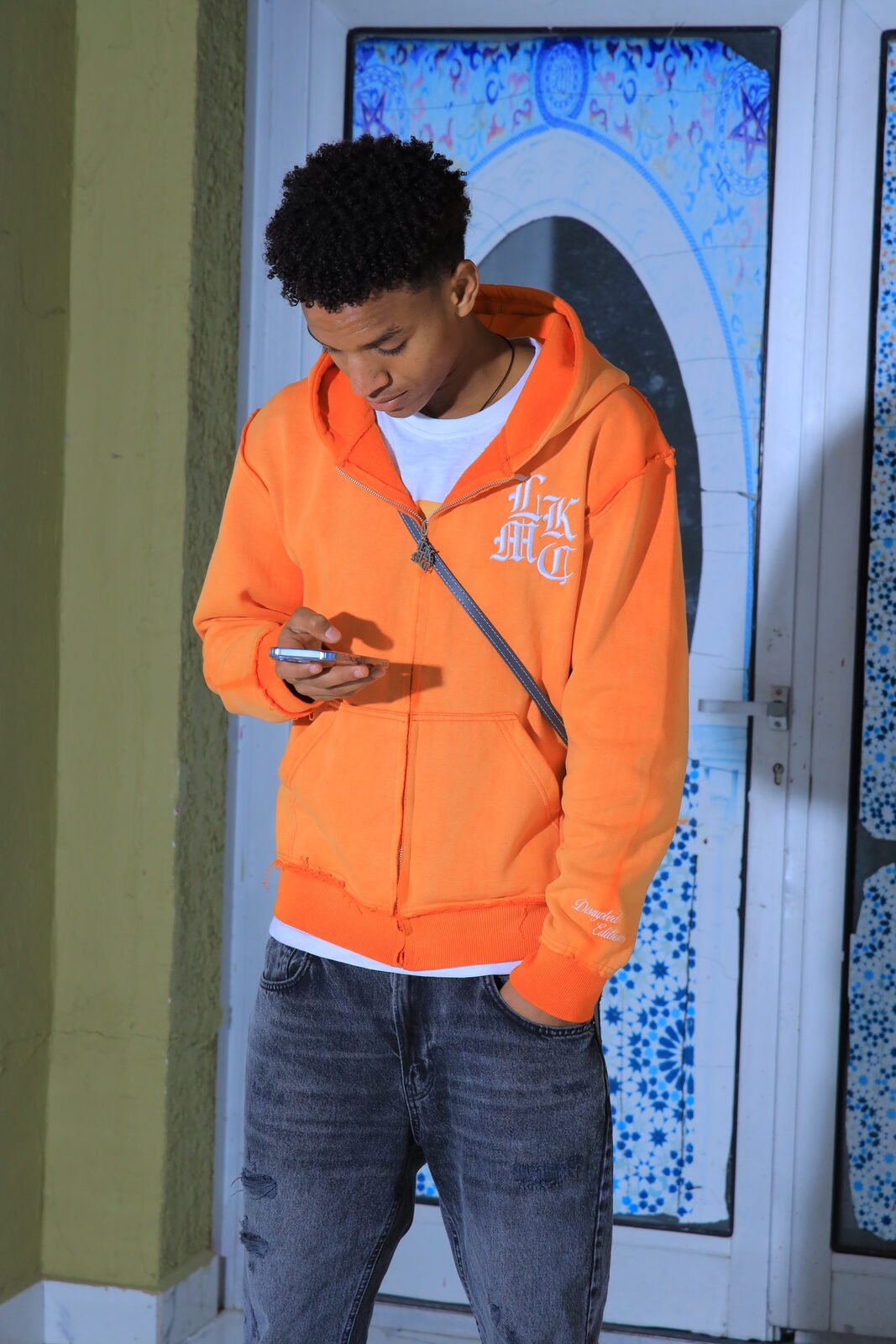Un jeune homme en sweat orange consulte son téléphone devant une porte à motifs bleus