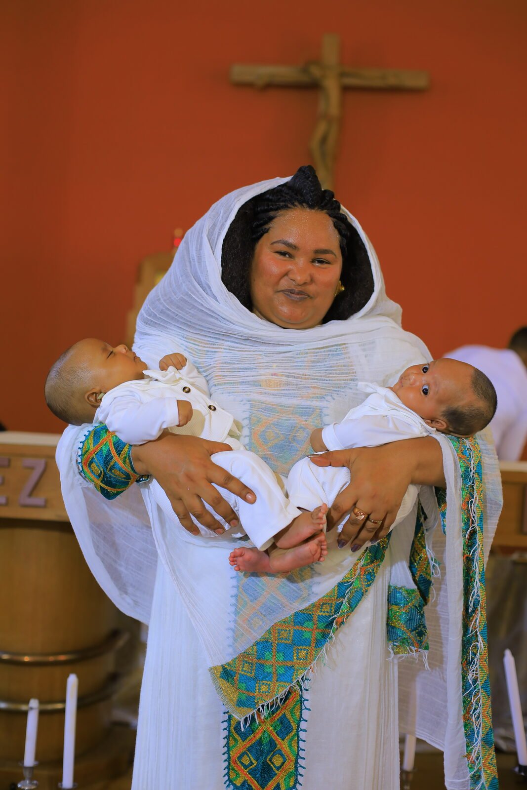 Une femme en tenue traditionnelle tient deux nourrissons, devant une croix
